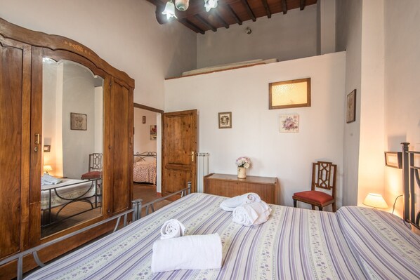 Un dormitorio con una cama, un armario de madera, un espejo, un escritorio, una silla y una mesita con un jarrón.
