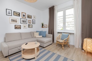 A modern living room with a sofa, a round coffee table, and a wicker chair. There are framed pictures on the wall and a window with curtains.
