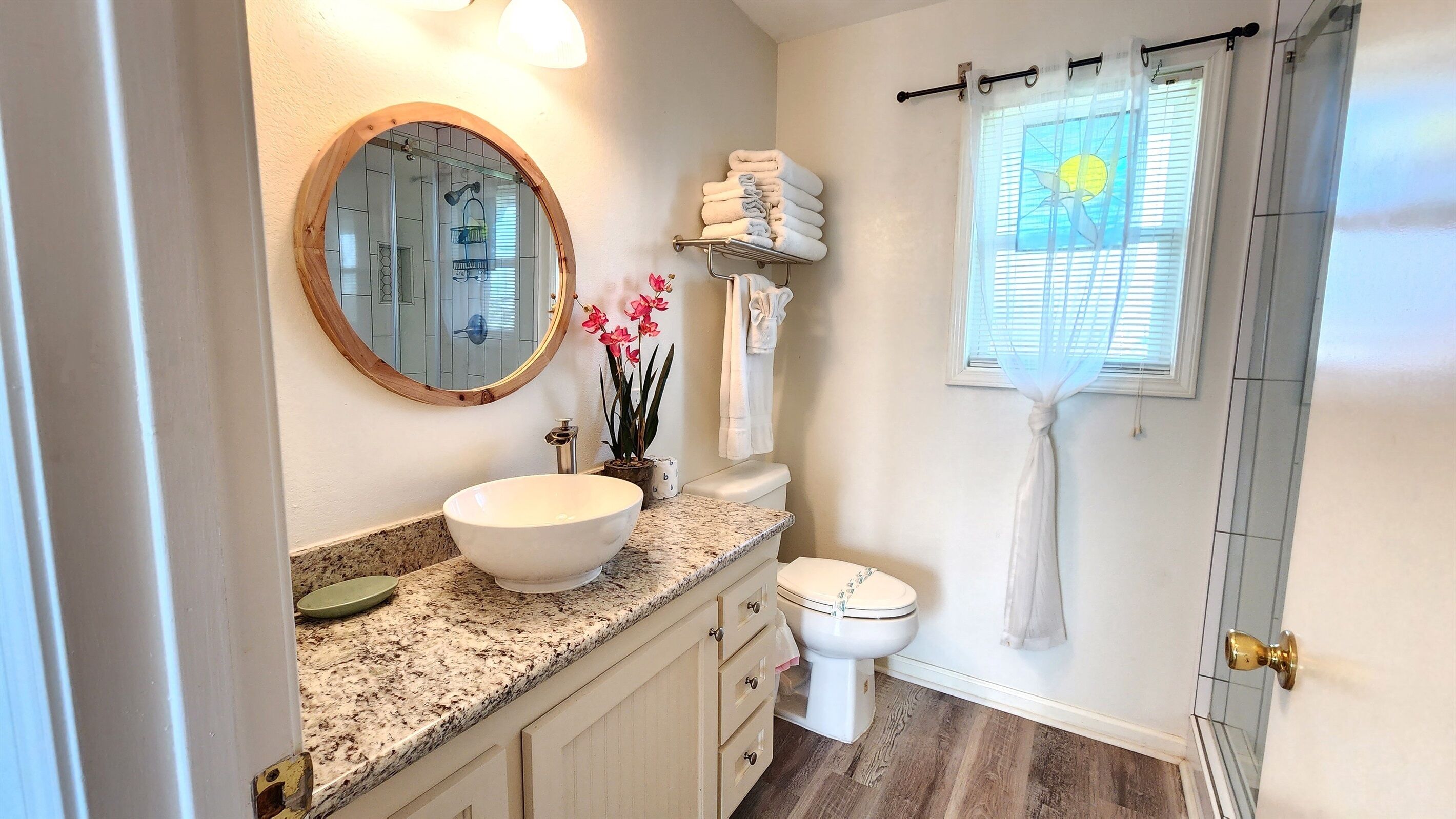 A bathroom with a round mirror, a sink, a toilet, and a window with a curtain.