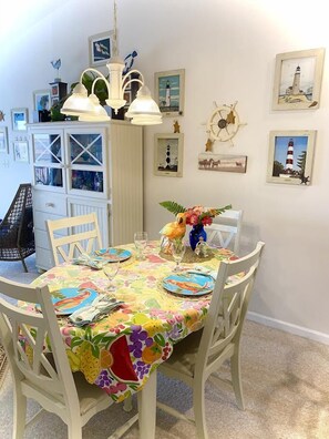 A dining area with a round table covered in a colorful tablecloth, white chairs, and a decorative bird centerpiece. - Cozy, comfortable 2 bedroom bayside condo with WiFi, an outdoor pool, and a breathtaking view of the bay located midtown and just a few blocks from the beach! (Ocean City)