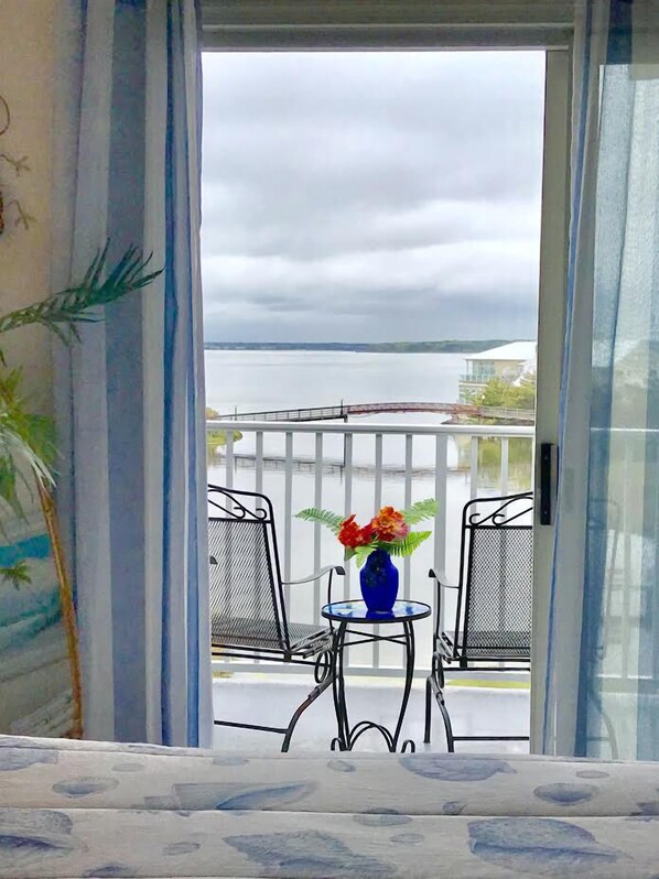 A balcony with a table and chairs, a blue vase with flowers, and a view of a body of water and distant buildings. - Cozy, comfortable 2 bedroom bayside condo with WiFi, an outdoor pool, and a breathtaking view of the bay located midtown and just a few blocks from the beach! (Ocean City)