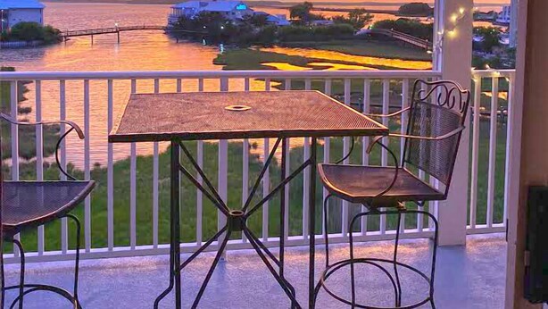 A balcony with a table and two chairs, overlooking a sunset over a body of water. - Cozy, comfortable 2 bedroom bayside condo with WiFi, an outdoor pool, and a breathtaking view of the bay located midtown and just a few blocks from the beach! (Ocean City)