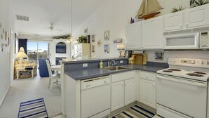 A kitchen with white cabinets, a microwave, stove, and a sink. There is a window with curtains, a chair, and a small table with a lamp. - Cozy, comfortable 2 bedroom bayside condo with WiFi, an outdoor pool, and a breathtaking view of the bay located midtown and just a few blocks from the beach! (Ocean City)