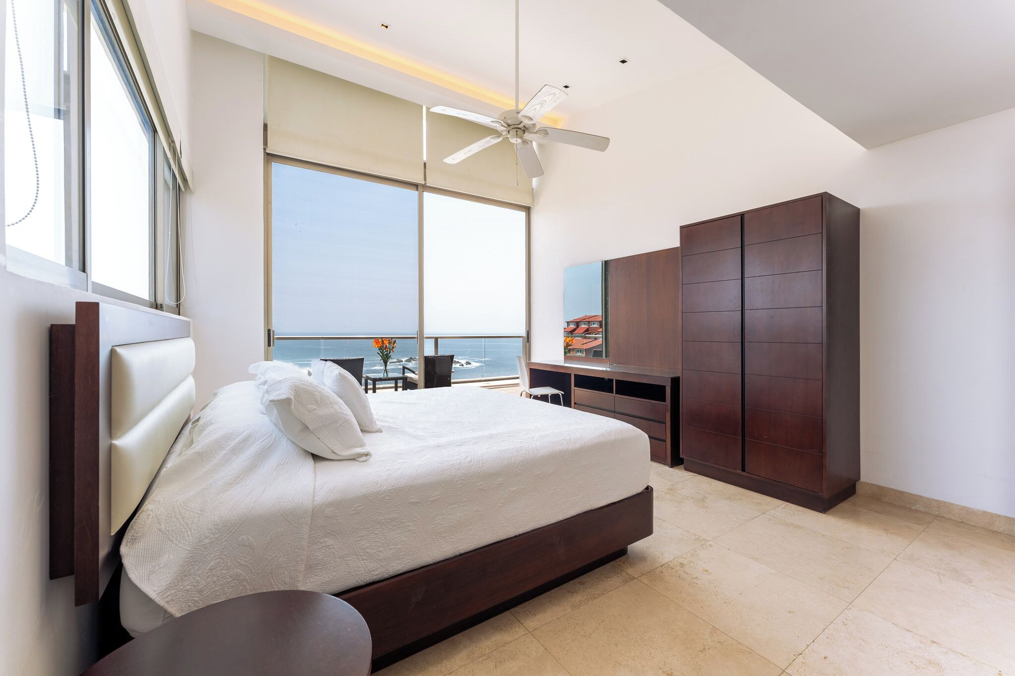 A modern bedroom with a large bed, a ceiling fan, a view of the ocean, and a large wardrobe.