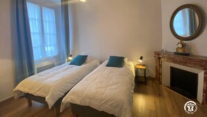 A bedroom with two beds, a fireplace, a mirror, and a window with curtains. - Agréable Maison en Pierre Avec Terrasse, au Coeur de la Citadelle (Villefranche-de-Conflent)