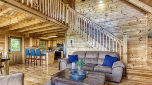 Interior de una cabaña de madera con una escalera, una sala de estar con sofá y sillones, y una zona de cocina.