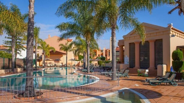 A resort pool area with palm trees, lounge chairs, and a building with columns.