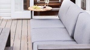 Eine geöffnete Tür führt zu einem Ziegelinnenraum, mit einem grauen Sofa, einem kleinen Tisch mit einem Weinglas und einem Laternen an der Wand.