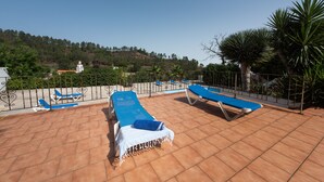 Eine Dachterrasse mit blauen Liegestühlen, einem weißen Tisch und Blick auf einen Hügel mit Bäumen und einem Gebäude.