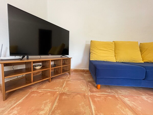 A living room with a flat-screen TV on a wooden stand, a blue sofa with yellow cushions, and a tiled floor. - Rural house, located in a quiet environment and surrounded by greenery (Fontanales (Moya))
