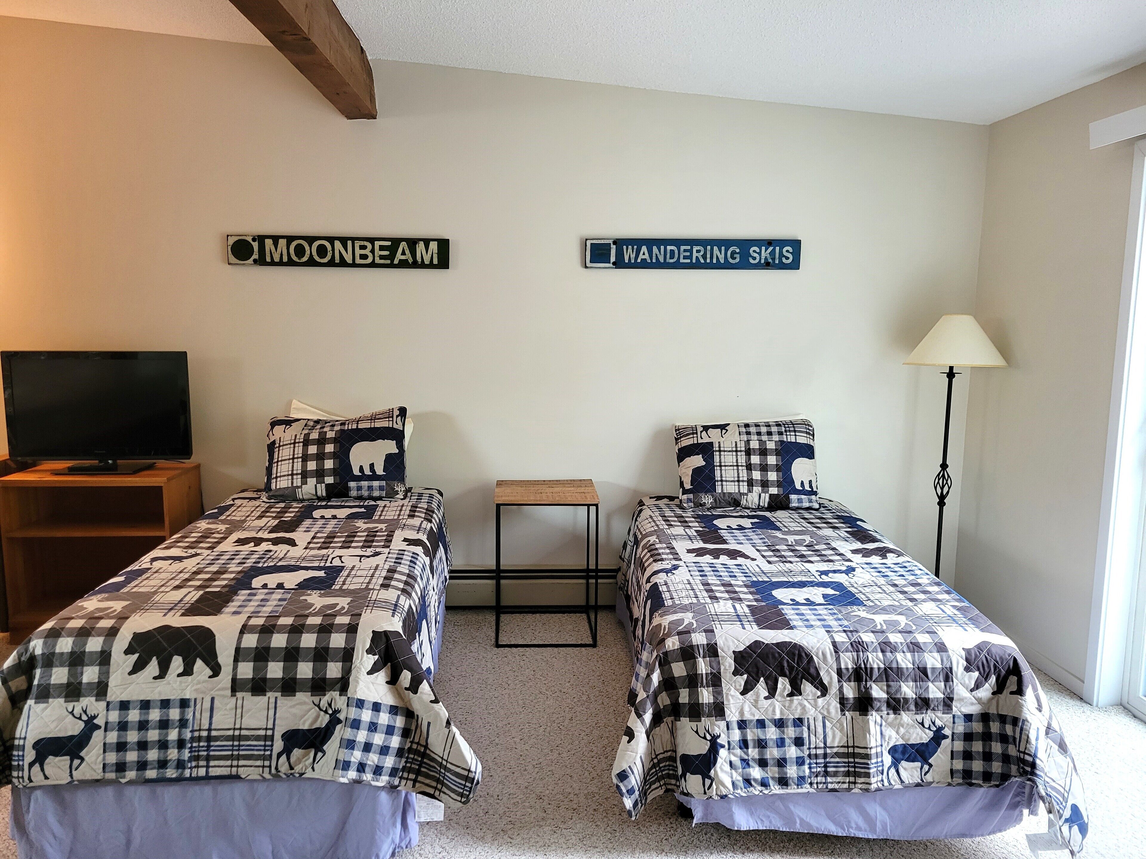 A bedroom with two beds, each with a checkered bedding featuring animal prints, a television on a wooden stand, and signs on the wall reading "MOONBEAM" and "WANDERING SKIS".