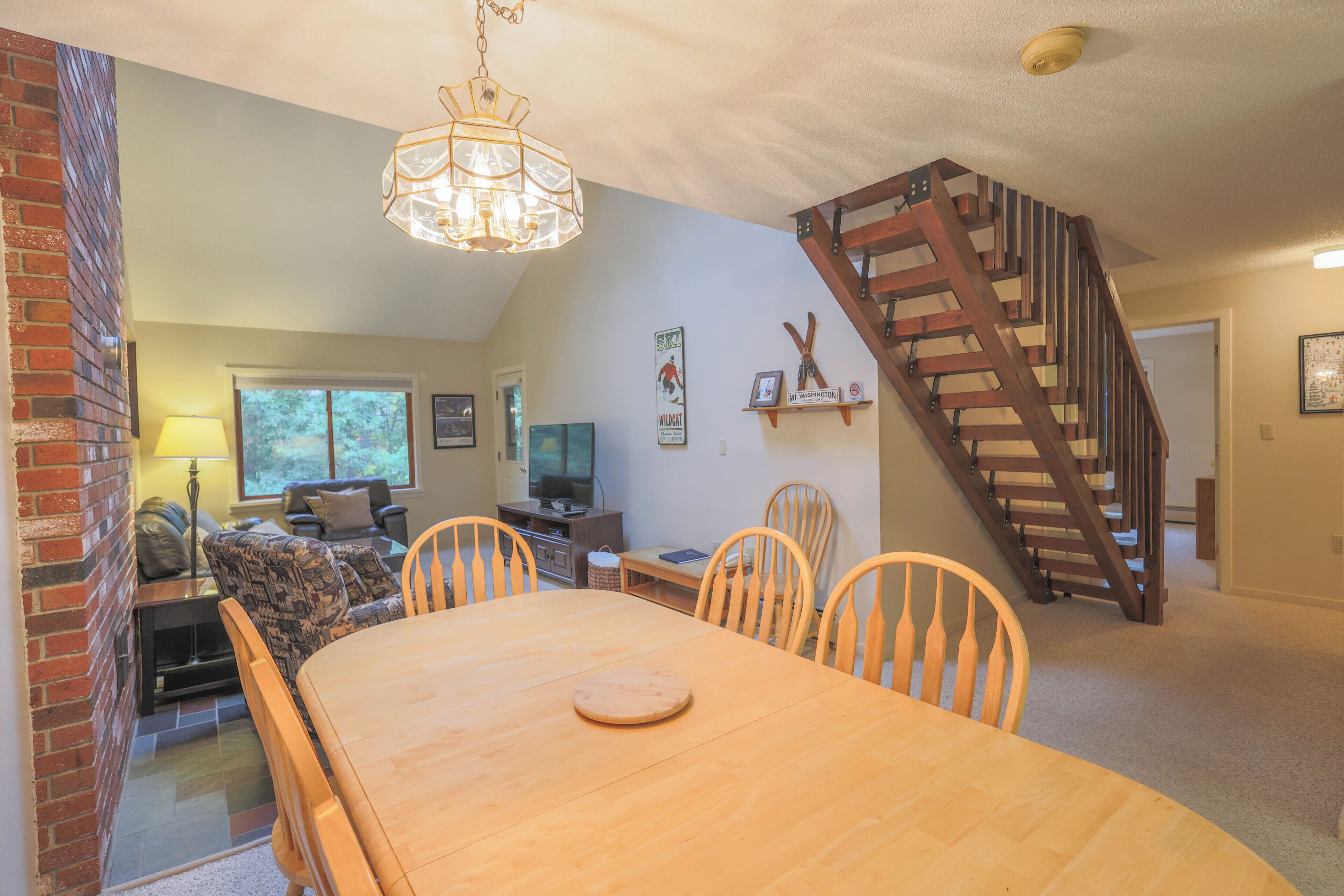A dining area with a wooden table and chairs, a brick wall, a fireplace, a television, and a wooden staircase.