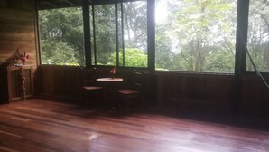 Habitación con suelo de madera, una mesa redonda con cuatro sillas y vistas a abundante follaje a través de ventanas amplias.