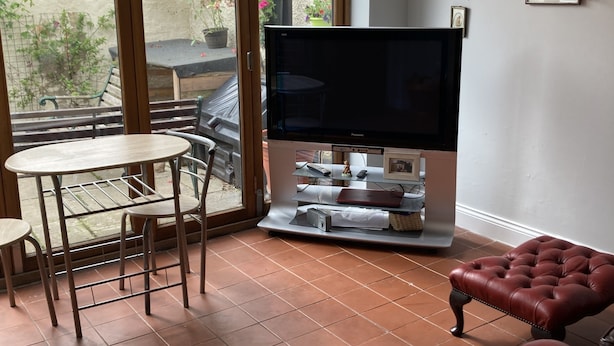 A living room with a television, a table with chairs, a cushioned bench, and a sliding glass door leading to an outdoor patio.