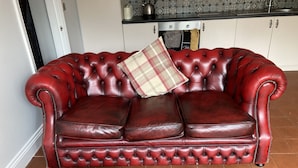 A red leather sofa with a plaid pillow in a room with a tiled floor and a kitchen in the background.