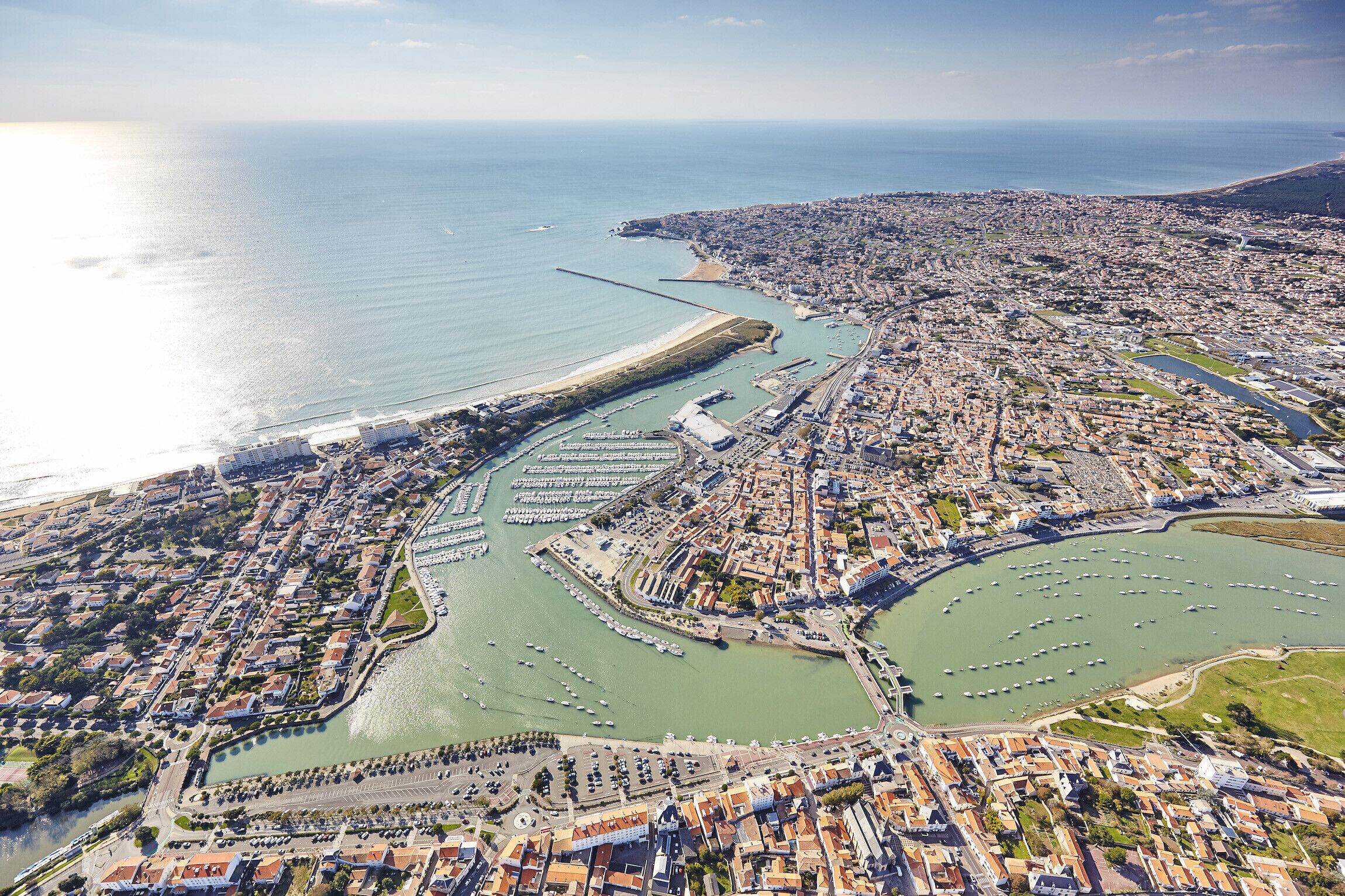Vista aérea de una ciudad costera con un puerto deportivo, zonas residenciales y una playa destacada.