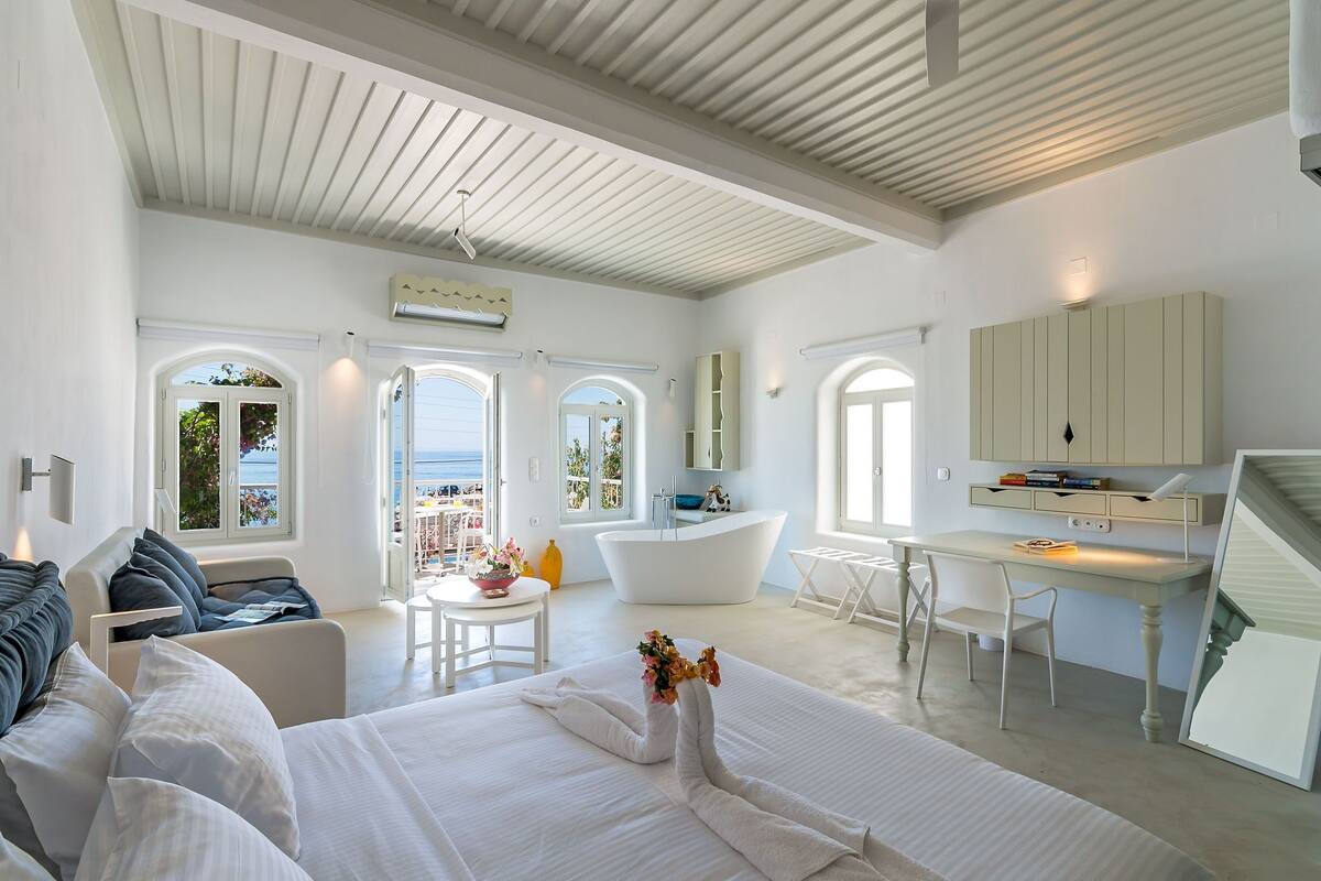 A modern bedroom with a large bed, a desk, and a bathtub. There are large windows and a view of the sea.