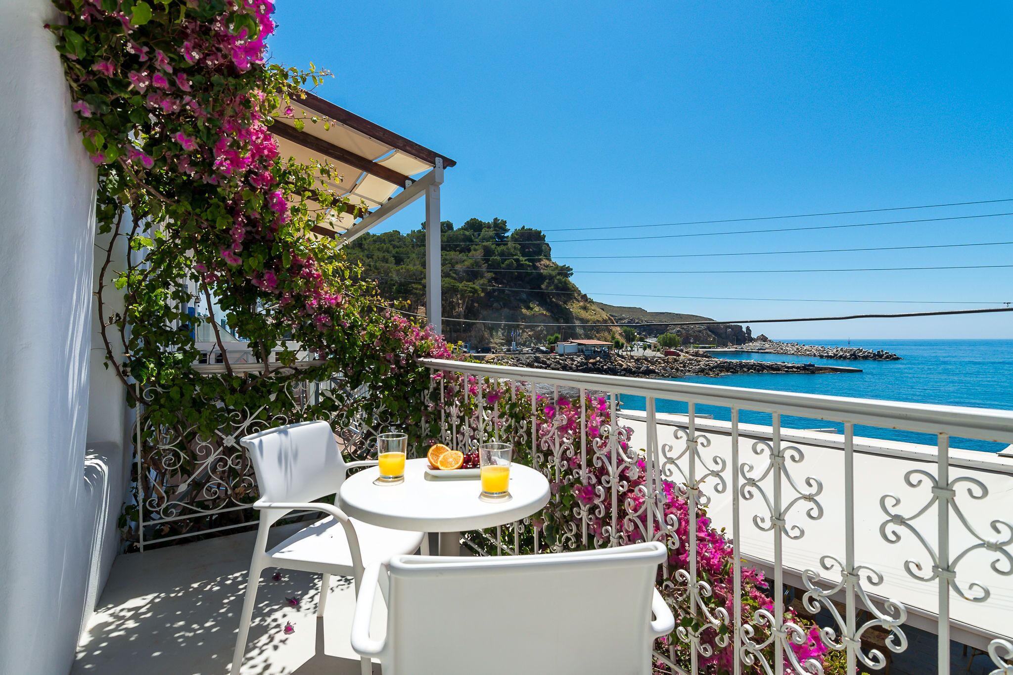 A balcony with white chairs and a table, overlooking a coastal view with a rocky shoreline and a clear blue sky.