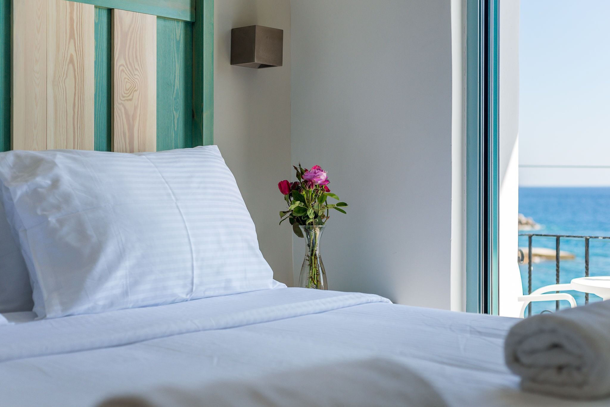 A neatly made bed with white linens, a vase with flowers, and a view of the sea through a balcony door.