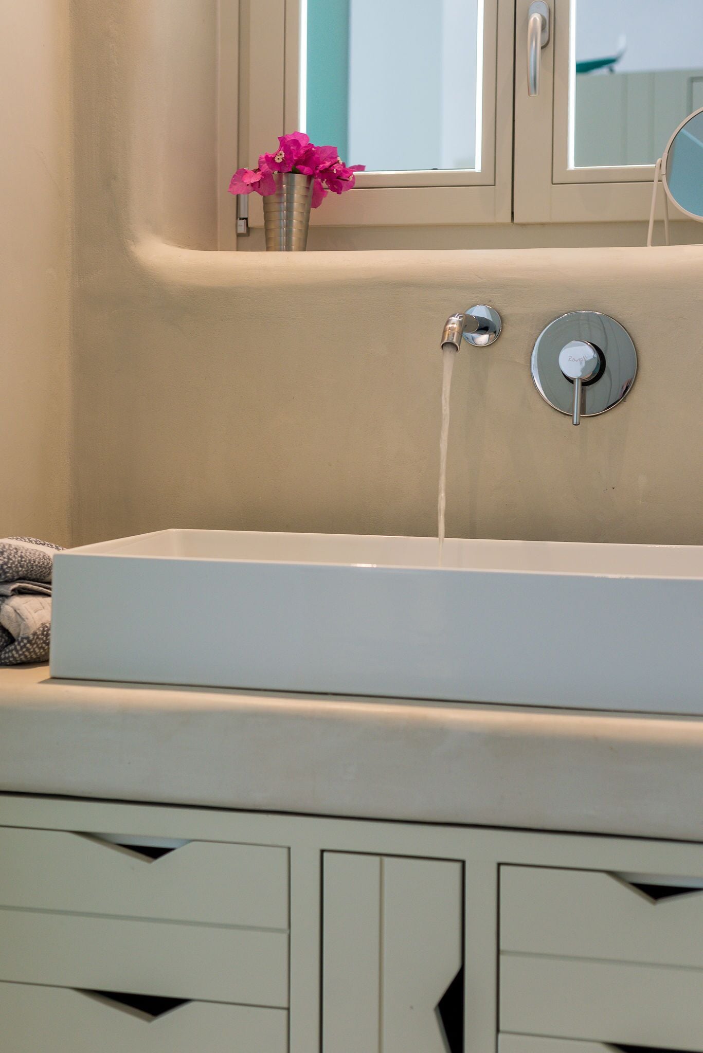 Un baño con un lavabo blanco, un espejo y una ventana con un jarrón de flores.