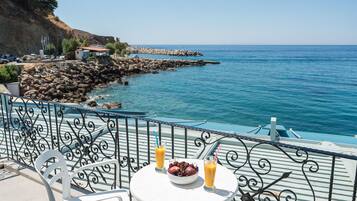 A table with a bowl of fruit and two glasses of orange juice on a balcony overlooking a rocky coastline and the sea.