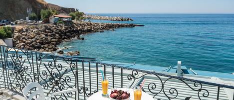 A table with a bowl of fruit and two glasses of orange juice on a balcony overlooking a rocky coastline and the sea.