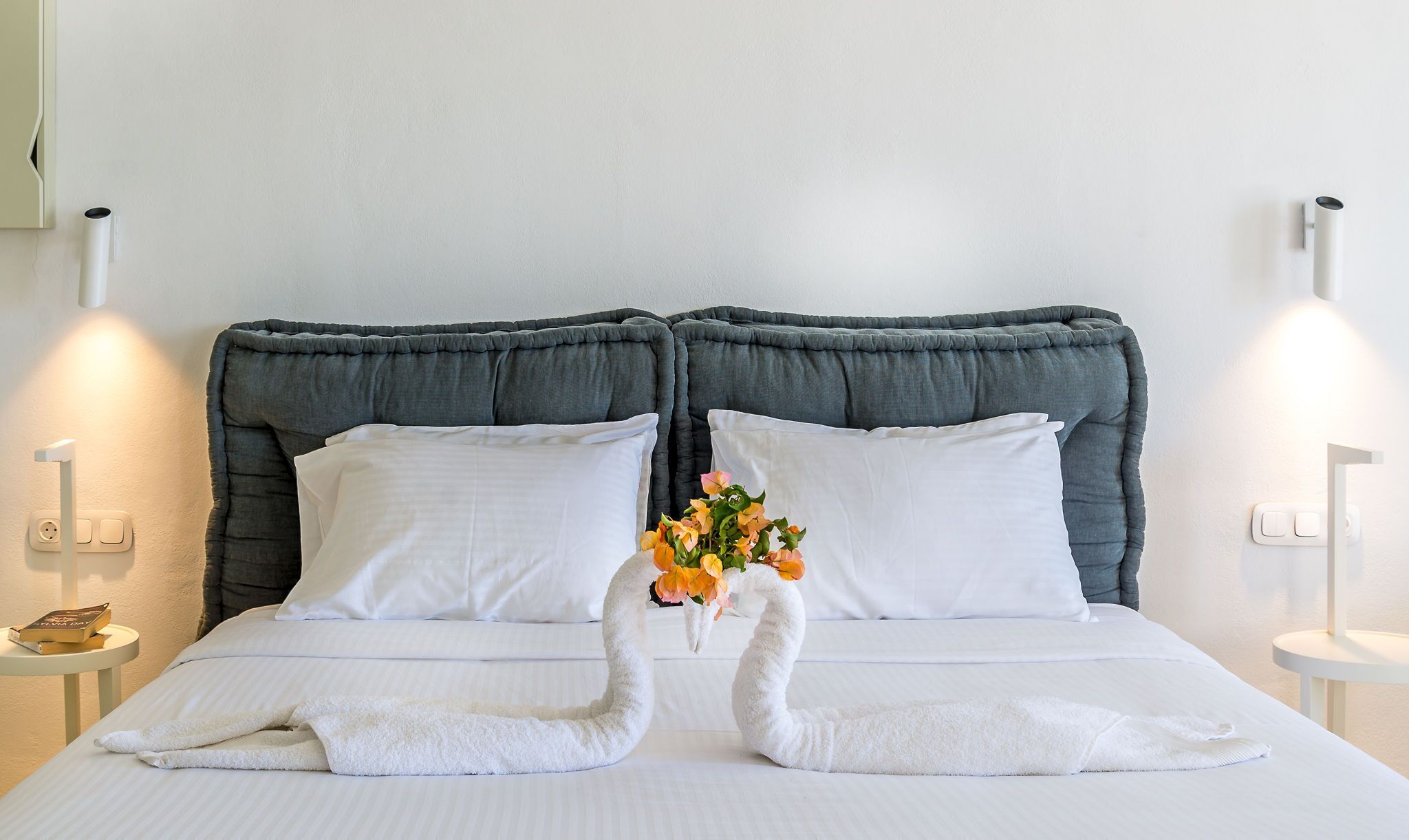 A bed with a grey upholstered headboard, white pillows, and a bouquet of flowers.