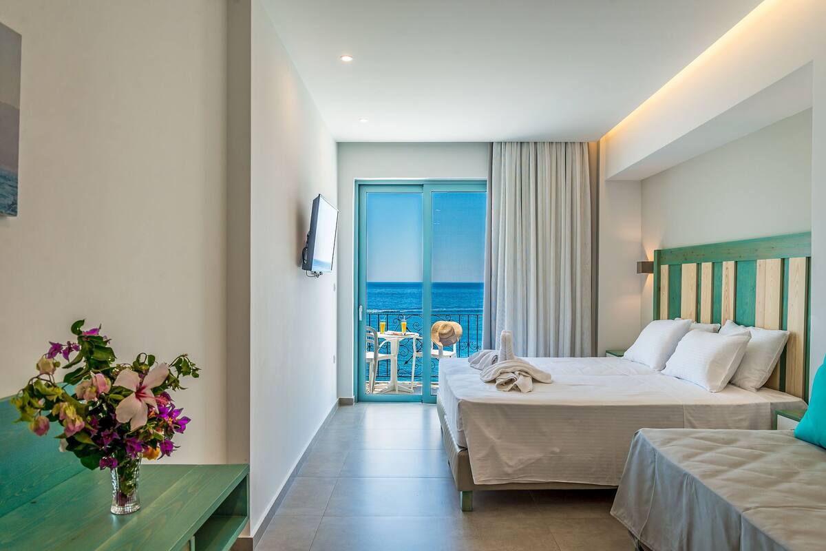 A hotel room with a bed, a small table with a chair, a TV, and a view of the sea.