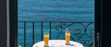A balcony with a view of the sea, two glasses of orange juice, a straw, a hat, and a newspaper on a white table.