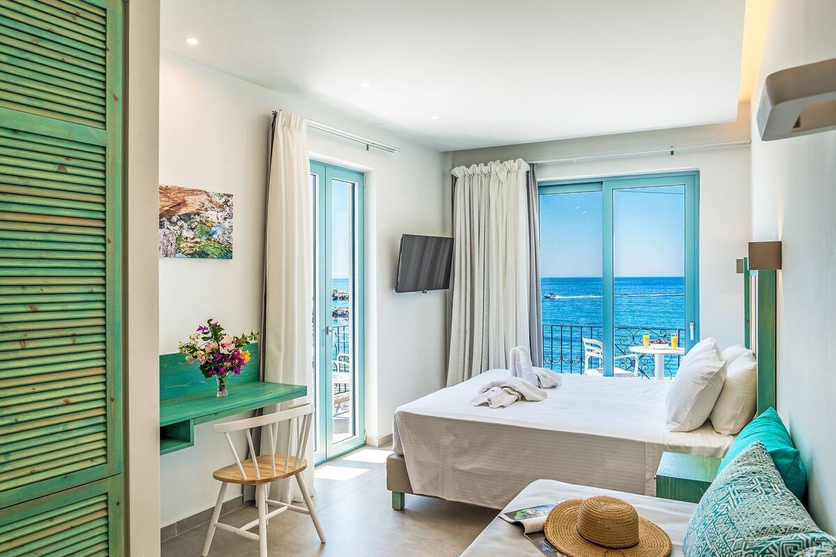 A bedroom with a bed, a desk, a chair, a TV, and a balcony with a view of the sea.