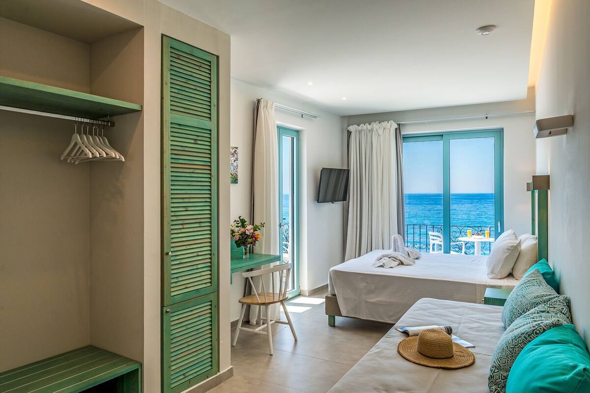 A bedroom with a bed, a desk, and a chair. There is a window with curtains, a TV mounted on the wall, and a view of the sea.