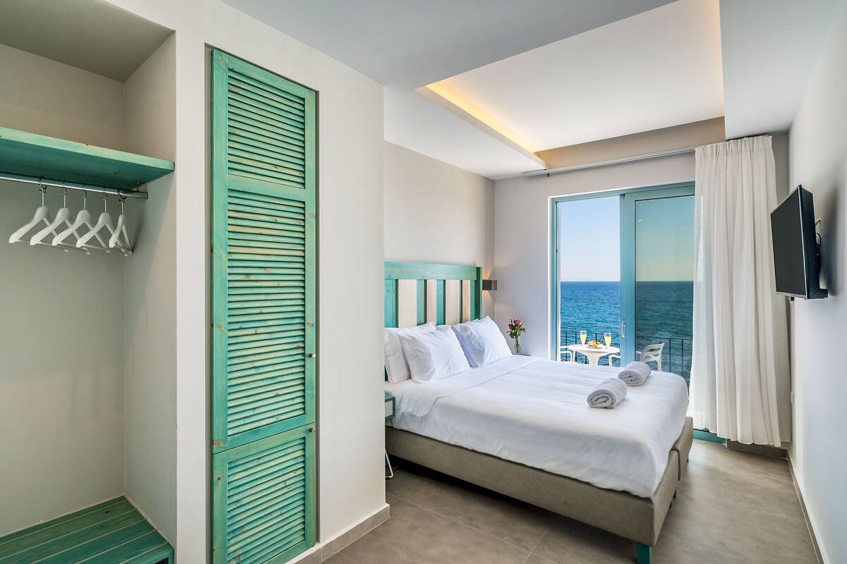 A hotel room with a bed, a TV, a balcony with a view of the sea, and a clothing rack.