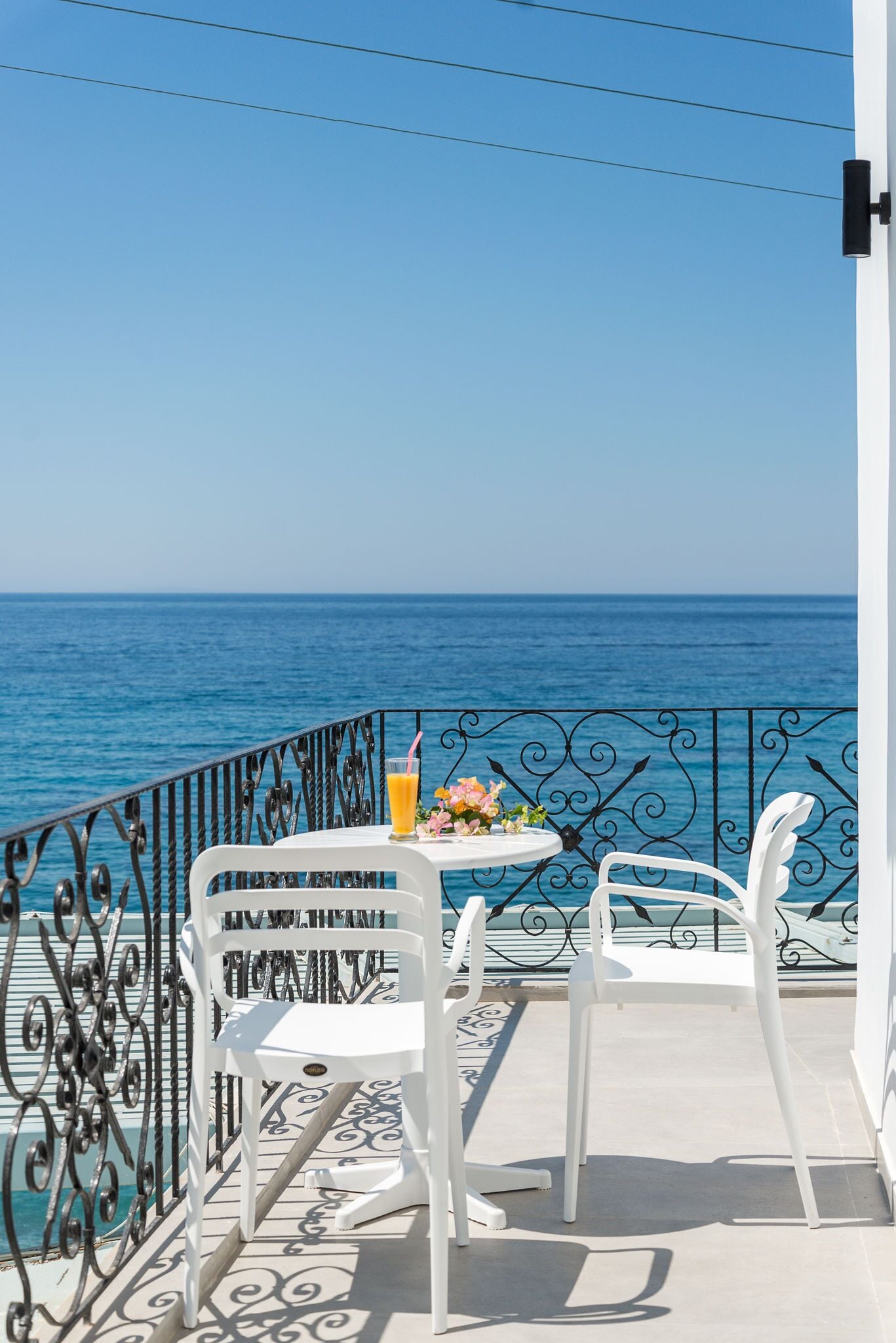 Un balcón con dos sillas blancas, una mesa con una bebida y flores, y vista al mar.
