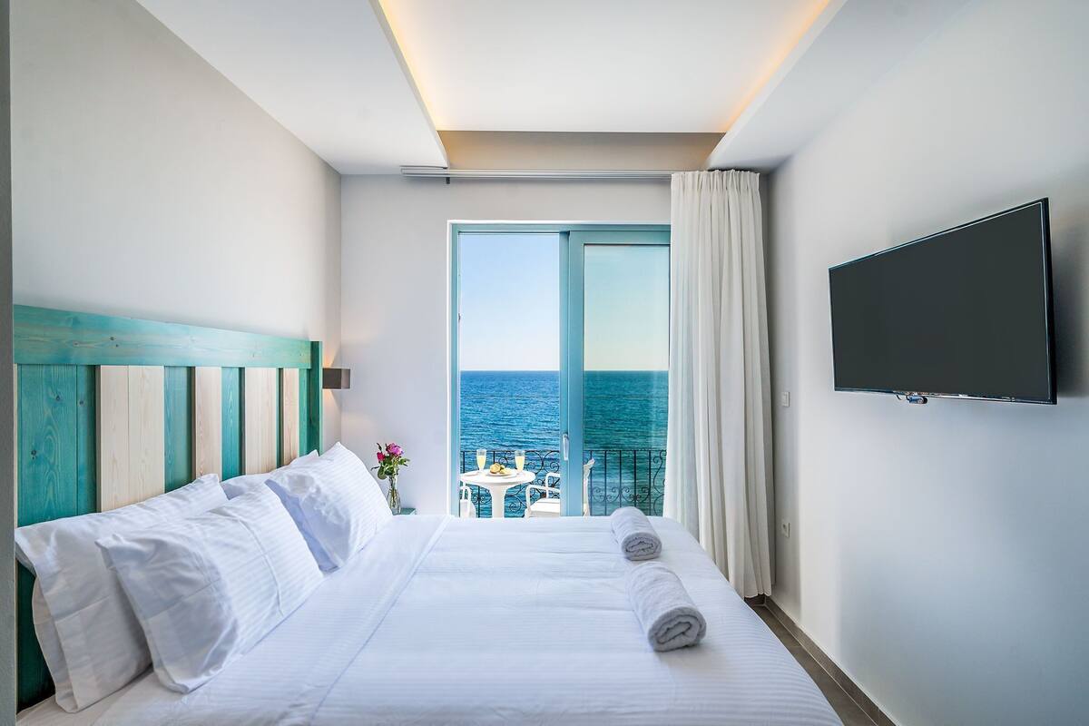 A hotel room with a bed, a TV, a balcony with a table and chairs, and a view of the sea.