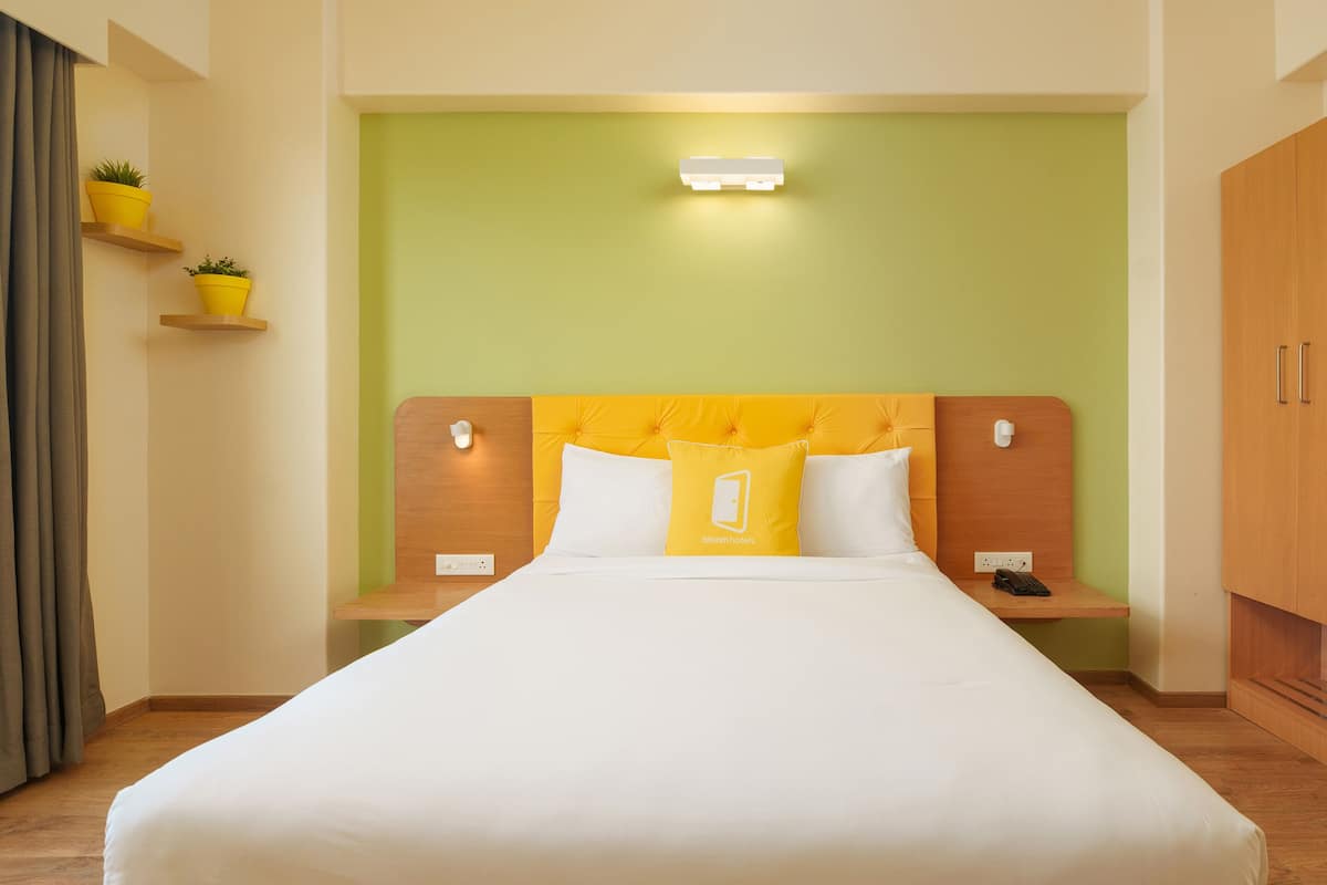 Habitación de hotel con cama, mesita de noche, lámpara, luz empotrada en la pared y una pequeña estantería con una planta.
