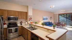 A modern kitchen with wooden cabinets, stainless steel appliances, a central island with a sink, and a dining area with a view. - Updated Condo with River Views! Near Alpine! (Alpine Meadows)