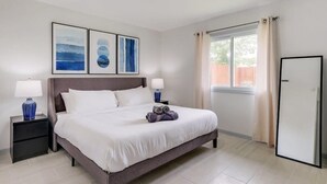Un dormitorio ordenado con una cama grande, dos mesitas de noche con lámparas, una ventana con cortinas y una puerta.