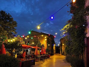 Ein Außenbereich zum Essen mit Tischen, Stühlen und Lichterketten, im Hintergrund ein Gebäude.