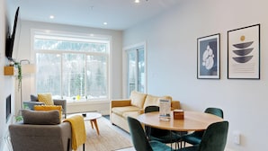 Una sala moderna con sofá, sillones, una mesa de centro y una mesa de comedor. Hay ventanas amplias con vistas a árboles y un edificio.