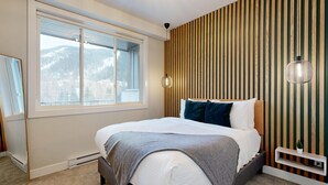 Un dormitorio moderno con una cama grande, una ventana con vistas a un paisaje nevado, un panel de pared de listones de madera y una lámpara fijada a la pared.