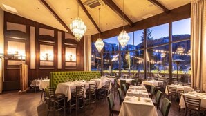 Un restaurante con una cabina tapizada de verde, varias mesas con manteles blancos, candelabros y amplias ventanas que dan a un paisaje nevado.