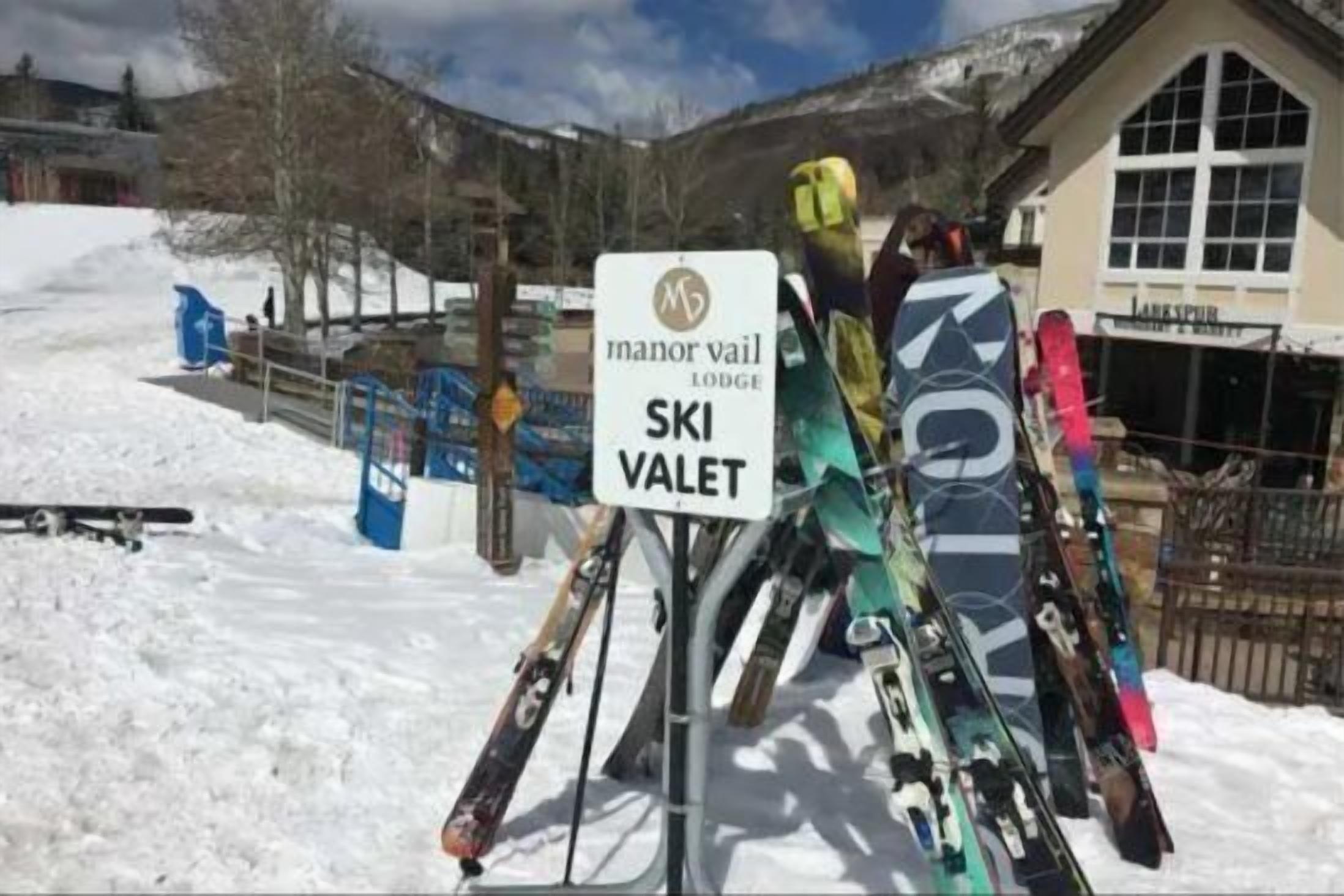 A ski resort sign with skis leaning against it, a mountain backdrop, and a lodge building.