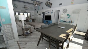 A spacious living room with a large wooden dining table, a comfortable sofa, and a television. - Kick's Kids - Channelfront in Garden City Beach! (Garden City Beach)