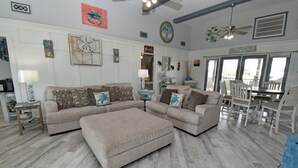 A spacious living room with a sectional sofa, a coffee table, and a dining area with white chairs and a table. - Kick's Kids - Channelfront in Garden City Beach! (Garden City Beach)