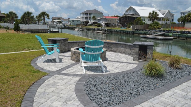 A waterfront patio with two Adirondack chairs, a stone fire pit, and a view of houses across the water. - Kick's Kids - Channelfront in Garden City Beach! (Garden City Beach)