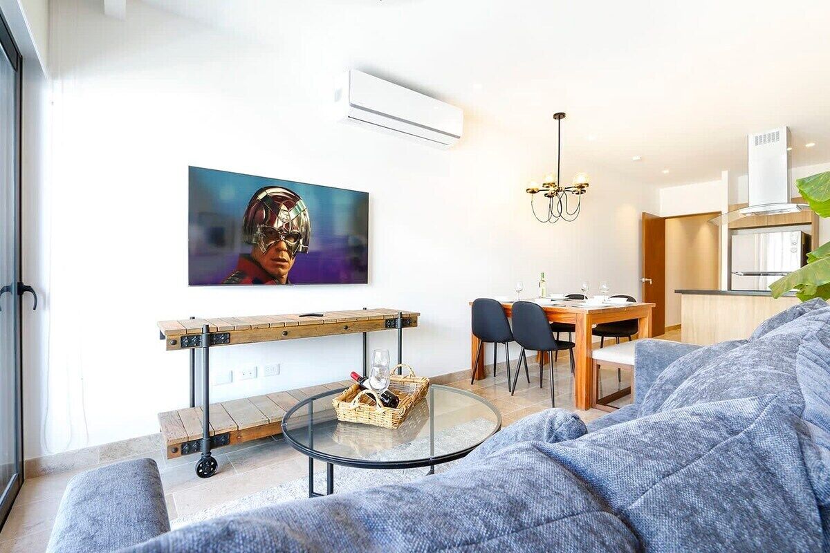Una sala de estar moderna con un sofá, una mesa de centro de vidrio, una zona de comedor con una mesa y sillas de madera, y un televisor montado en la pared.