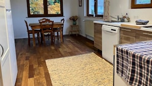 Une cuisine avec un plancher en bois, une table à manger avec des chaises, une fenêtre donnant sur un paysage automnal et un lave-vaisselle.