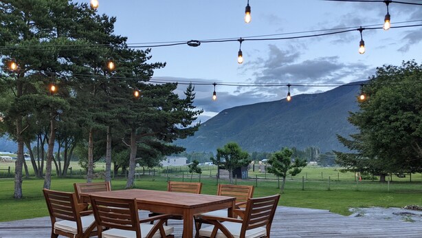 A wooden deck with a table and chairs, surrounded by trees and a mountainous landscape. - 3/2 home 20 minutes from Glacier National Park, Sleeps 8! (Columbia Falls)