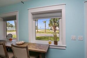 A room with a dining table, chairs, a window with a view of palm trees and a road, and a welcome sign. - 175 Steps from the Beach, Sweeping Views, Ground Floor (Mexico Beach)