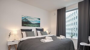 Una habitación de hotel moderna con una cama grande, una mesita de noche con lámpara, vistas a un paisaje urbano a través de la ventana y un cuadro en la pared.